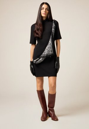 Vestido negro de manga corta con una bufanda de patrón diagonal en blanco y negro. La modelo lleva guantes de cuero negros y botas marrones hasta la rodilla.