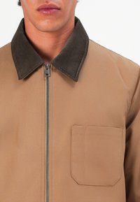 Brown zip-up jacket featuring a dark brown corduroy collar and a single chest pocket with a rounded bottom edge. Smooth fabric texture.
