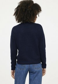Dunkelblaues gestricktes Sweatshirt mit langen Ärmeln und rundem Ausschnitt, mit einem gerippten Saum. Trägt dazu eine hellblaue Jeans.