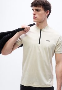 Νέος άνδρας που κοιτάζει προς τα δεξιά, φοράει μπεζ κοντομάνικο πουκάμισο με φερμουάρ και το κείμενο "BOSS", κρατώντας ένα μαύρο σακάκι στον ώμο του.