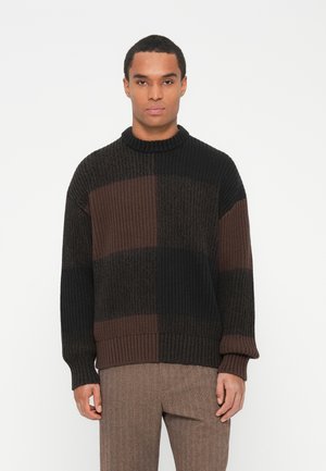 Vīrietis ar ribotu, krāsu blokētu džemperi tumši brūnā un melnā krāsā, valkājot brūnas herringbone bikses, stāv pretī vienkāršai gaišai fona.