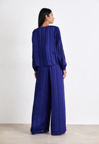Ensemble en deux pièces dans une teinte bleu profond, avec des rayures verticales. Le haut a une coupe loose avec des manches longues ; le pantalon à jambe large tombe élégamment.