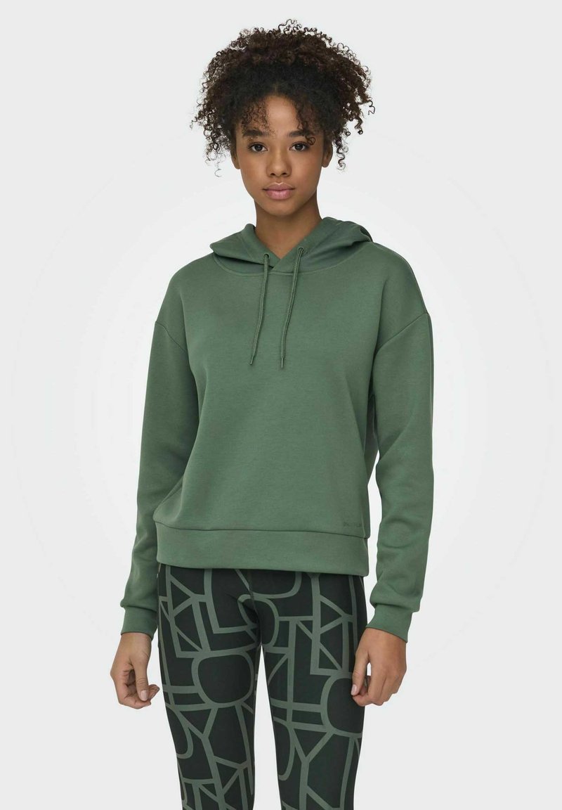 Grüne Kapuzenjacke mit Kordelzug, entspannter Schnitt und gerippten Bündchen. Kombiniert mit gemusterten schwarzen Leggings mit geometrischen Designs.