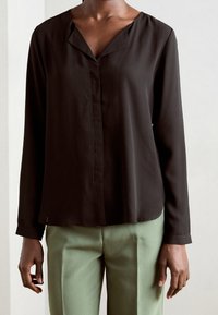Blusa nera a maniche lunghe con scollatura a V, realizzata in tessuto liscio. Presenta una piastra frontale e una vestibilità leggermente svasata, abbinata a pantaloni verde chiaro.