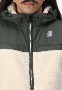 Giacca imbottita verde con pannello in pile crema sulla parte frontale, dotata di chiusura con zip, cappuccio e logo sul lato sinistro superiore.