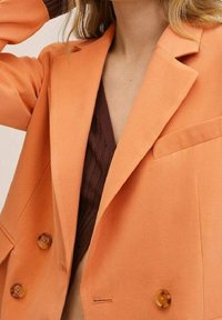 Blazer orange à double boutonnage avec revers crantés, doté de grands boutons ambre et d'une texture de tissu lisse, porté sur un haut foncé.