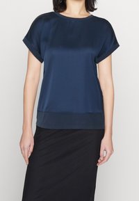 Top blu navy a maniche corte in tessuto liscio con scollatura rotonda e maniche arrotolate, caratterizzato da un orlo scuro a contrasto e una silhouette elegante.