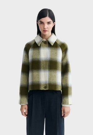 Jeune femme aux cheveux raides et foncés portant une grande veste en laine à carreaux verts et blancs sur un pantalon noir, debout devant un fond clair uni.