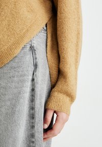 Person wearing a textured tan sweater with sleeves covering wrists and light gray denim jeans, hand relaxed by the side against a plain background.