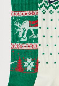Zelená ponožka s bílými soby, červeným sane a sněhovými vločkami, vedle bílé ponožky s zeleným diamantovým a tečkovaným vzorem.