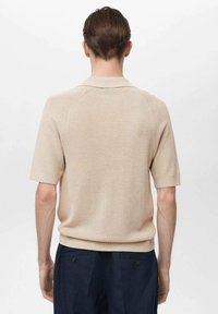 Béžová pletená polo trička s krátkým rukávem a texturovaným povrchem, s žebrovaným límcem a volným střihem, spárované s tmavými kalhotami.