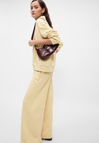 Ανοιχτό κίτρινο υπερμεγέθη φούτερ και παντελόνια με φαρδιά πόδια, συνδυασμένα με μια μπορντό καμπυλωτή τσάντα χειρός με λεπτομέρεια από κούμπωμα. Λείο υλικό.