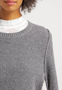 Grå stickad tröja med rund halsringning, prydd med ett strukturerat stickmönster och synliga sömmar, lagd över en vit spetsblus.
