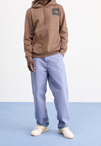 Osoba oblečená v hnědém mikině The North Face, světlých modrých volných kalhotách a krémových teniskách stojí na bledě modré podlaze proti bílé zdi.