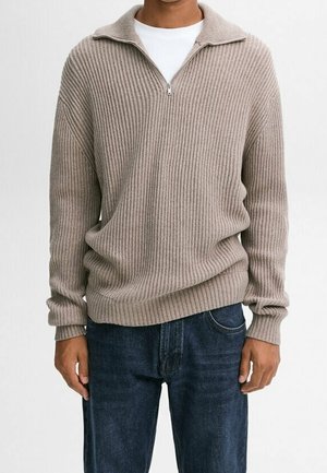 Man die een beige gebreide trui met kwartritskraag draagt over een wit overhemd en donkerblauwe spijkerbroek, staand tegen een effen achtergrond.