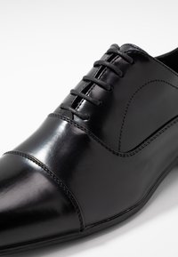 Chaussure habillée en cuir noir avec une surface lisse et élégante, un bout pointu et un laçage traditionnel. Coutures détaillées sur les côtés.