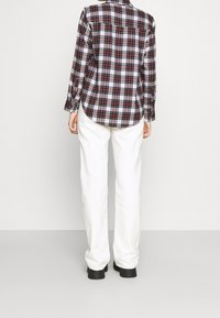 Geruit flanellen shirt in rood en zwart met een gebogen zoom, gecombineerd met witte wijde broek en zwarte haklaarzen. Neutrale achtergrond.