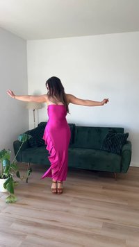 Fekvő magenta selyemruha aszimmetrikus fodrokkal az oldalon, bézs platform szandálokkal kombinálva, egy minimalista szobában zöld kanapéval.