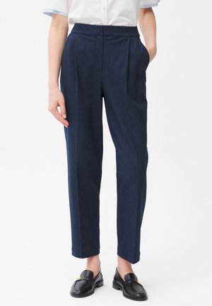 Kobieta nosząca granatowe spodnie o wysokim stanie, zwężane z kieszeniami po bokach, do tego czarne mokasyny i biała koszula z krótkim rękawem.