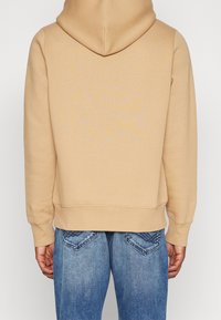 Felpa beige con cappuccio a coulisse, tessuto morbido e polsini a coste. Abbinata a jeans blu con tasche posteriori decorative.