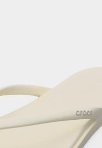 Μπεζ σανδάλι τύπου φλιπ-φλοπ Crocs με λεπτά λουράκια και ανάγλυφο λογότυπο Crocs στο πλάι, σε λευκό φόντο.