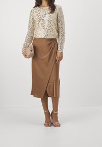 Zlatý dlouhý rukáv se třpytkami v kombinaci s hnědou omotanou sukní, držící béžovou kabelku a v béžových sandálech na vyšším podpatku.