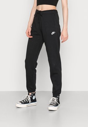 Osoba ubrana w czarne spodnie dresowe Nike z sznurkiem w talii oraz czarne wysokie trampki z białymi sznurówkami i podeszwami, stojąca na jednolitym tle.