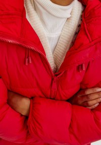 Červená péřová bunda s vysokým límcem a dvojitými zipy, navrstvená přes bílý žebrovaný svetr, která předvádí měkkou, zateplenou texturu.