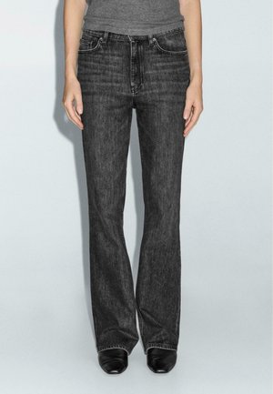 Persoon die donkergrijze, recht gesneden jeans met hoge taille en zwarte schoenen draagt, staand tegen een effen lichte achtergrond.