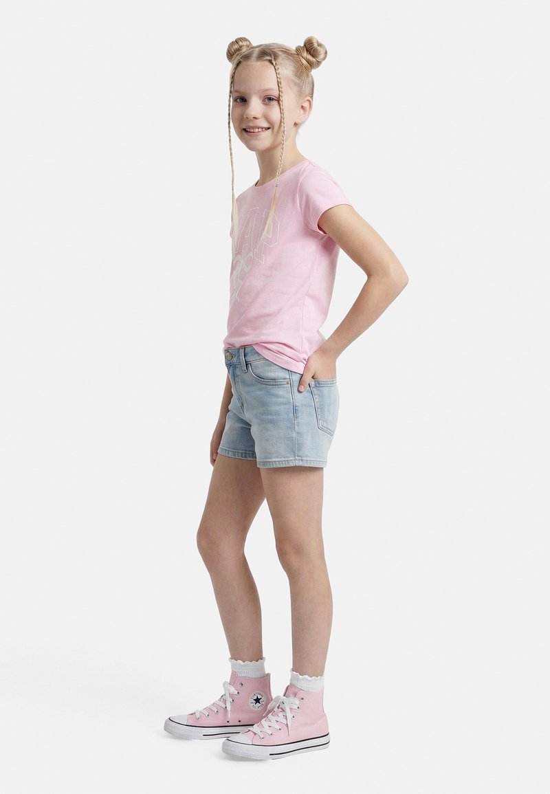 Νεαρή κοπέλα με ξανθά μαλλιά σε δύο κότσους, φοράει ροζ μπλούζα, τζιν σορτς, λευκές κάλτσες και ροζ ψηλά αθλητικά παπούτσια, στέκεται πλαγιομεετρημένη και χαμογελάει.