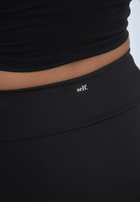Crni sportski top s prilagođenim dizajnom i glatkom teksturom. Ima mali bijeli logotip "self." na struku.