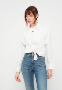 Donna con capelli rossi che indossa una camicia bianca con bottoni annodata in vita sopra una maglietta bianca e jeans a vita alta blu, su uno sfondo bianco uniforme.
