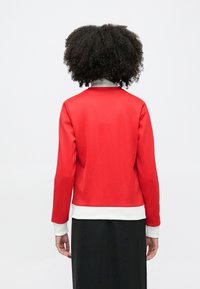 Persona ar cirtainiem matiem, valkājot sarkanu garroku kreklu ar baltiem manžetiem un apakšmalu, stāv pret vienkāršu balto fonu, ar muguru pret kameru.