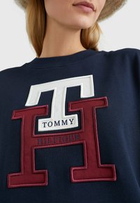 Námořnická modrá mikina s velkým vyšívaným logem v bílé a burgundské barvě s textem "TOMMY HILFIGER" níže, žebrovaným kulatým výstřihem.