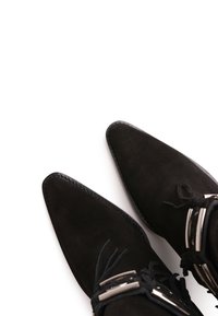 Botas negras de ante hasta el tobillo con puntas afiladas, cordones decorativos y detalles de hardware plateado. Textura suave con un borde cosido.
