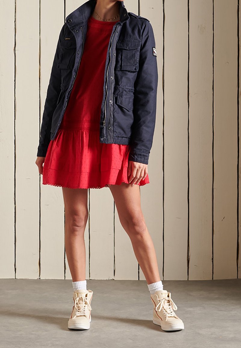 Personne portant une robe rouge, une veste bleu marine avec des poches, des chaussettes blanches et des baskets montantes beiges, debout devant un fond en panneaux de bois blancs.