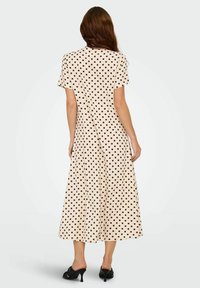 Beige midi-klänning med svarta prickar, korta ärmar, en lös passform och en utsvängd fåll. Matchas med svarta högklackade skor.