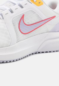 Scarpa sportiva bianca con tomaia testurizzata, con il logo Nike in rosa e viola, e una suola grigia spessa con scanalature di trazione.