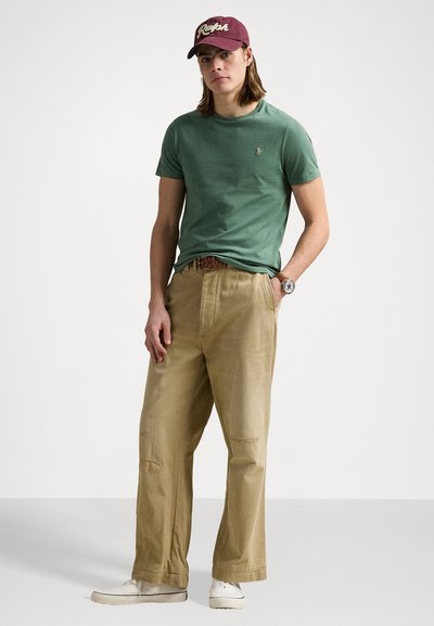 Green cotton t-shirt with a logo, beige relaxed-fit pants with rips, white shoes, and a maroon cap with light lettering.