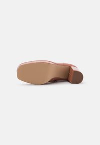 Zapato de tacón de cuero marrón tumbado de lado, mostrando una puntera cuadrada y un tacón cuadrado sobre un fondo blanco liso.