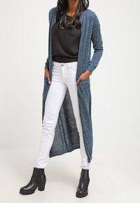 Cardigan lungo di un blu scuro con apertura frontale e maniche lunghe, abbinato a una maglietta nera e jeans bianchi attillati, indossato con stivali con tacco neri.