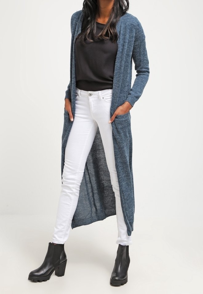 Cardigan lungo di un blu scuro con apertura frontale e maniche lunghe, abbinato a una maglietta nera e jeans bianchi attillati, indossato con stivali con tacco neri.