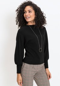 Maglione nero a maglia con collo alto e maniche a sbuffo. Abbinato a pantaloni a quadri. Collana lunga d'argento con un pendente circolare.