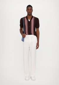 Hnědá košile s krátkým rukávem a růžovými vertikálními pruhy, v kombinaci s bílými kalhotami na stažení a bílými teniskami. Na rameni je modrá taška.