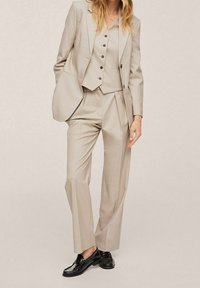 Completo beige su misura composto da blazer aderente, gilet coordinato con bottoni e pantaloni a gamba larga, completato da scarpe nere.