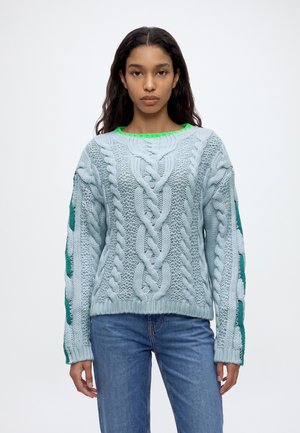 Un mannequin se tient devant un fond blanc uni, vêtu d’un pull bleu clair à torsades, d’un jean bleu et de baskets, et portant un sac à main marron.