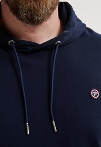 Felpa blu navy con tasca frontale, cordini regolabili con punte in metallo e un piccolo logo circolare sul petto.