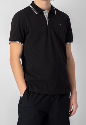 Uomo che indossa una polo nera con bordi bianchi su colletto e maniche, in piedi con le mani nelle tasche dei pantaloni neri contro uno sfondo semplice.