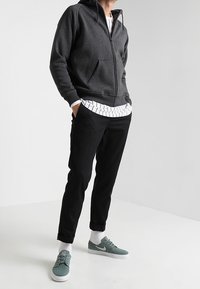 Sudadera con cremallera de color gris oscuro y bolsillo tipo canguro, camiseta de rayas blanca por debajo, pantalones negros y zapatillas verdes con suelas blancas.
