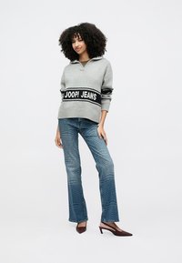 Grauer Pullover mit schwarzem Band und der Aufschrift "JOOP! JEANS", kombiniert mit blauen Schlagjeans mit Strukturmuster und bordeauxfarbenen spitzen Schuhen.